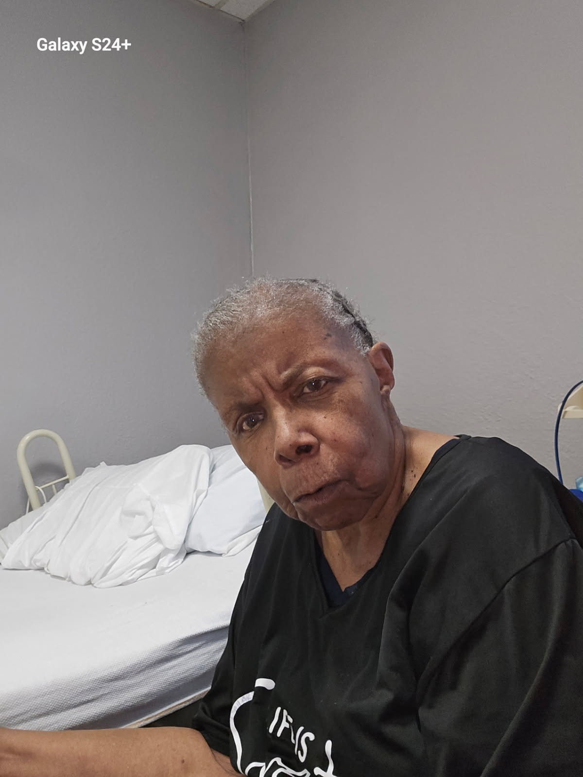 The same woman sitting beside a bed in a plain gray care-facility room, looking at the camera with a stern, skeptical expression