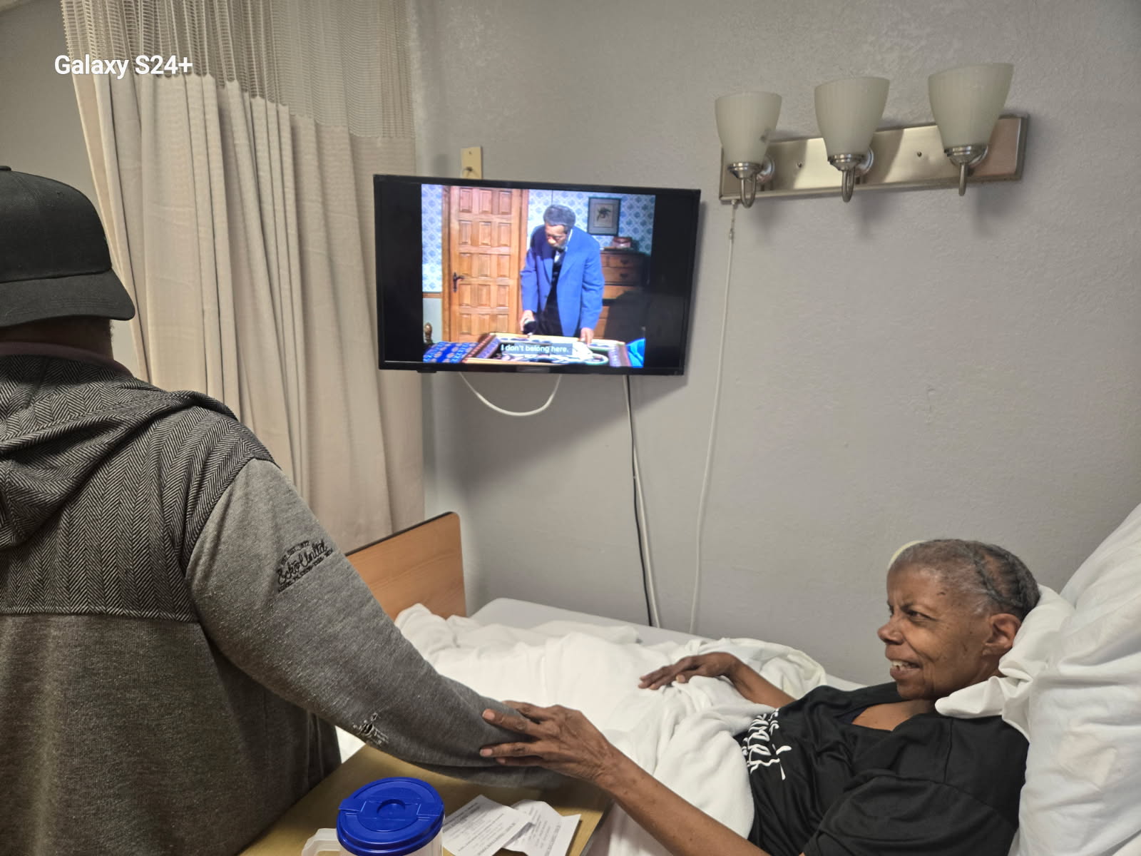 The woman resting in bed, smiling and holding the arm of a man in a gray hoodie and cap standing beside her, with a TV on the wall
