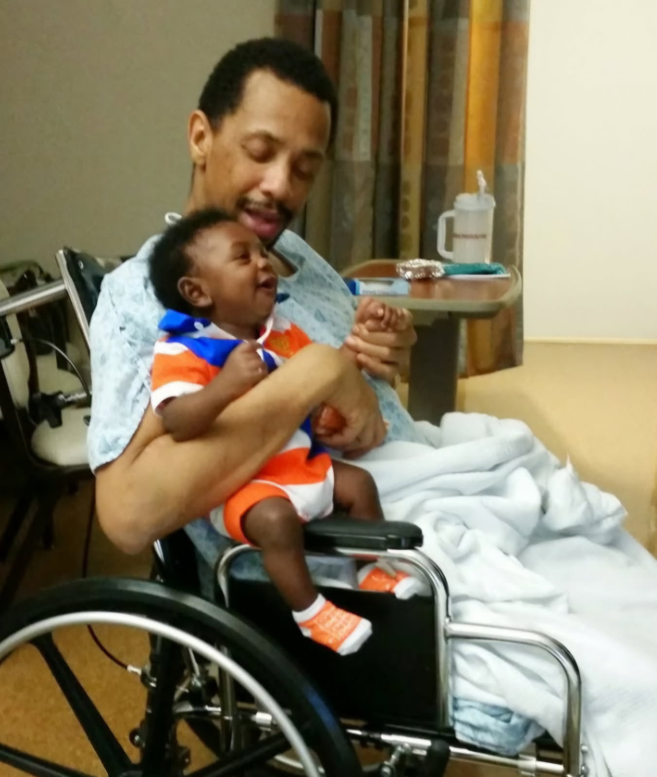 A man in a wheelchair and hospital gown smiling while holding a laughing baby