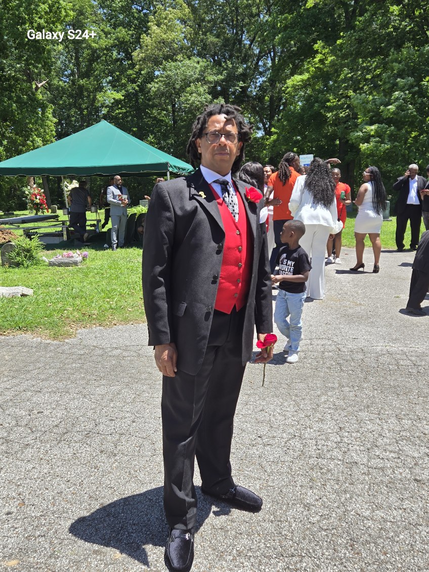 A man in a dark suit and red vest standing outdoors at a family gathering holding a rose