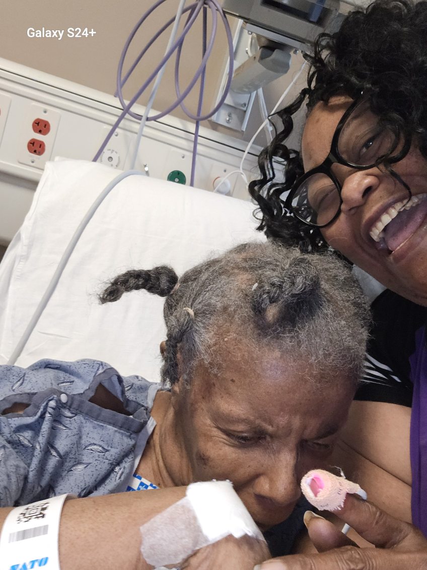 An older woman in a hospital gown lying in a hospital bed with braided gray hair and a wristband, while our founder leans in beside her smiling