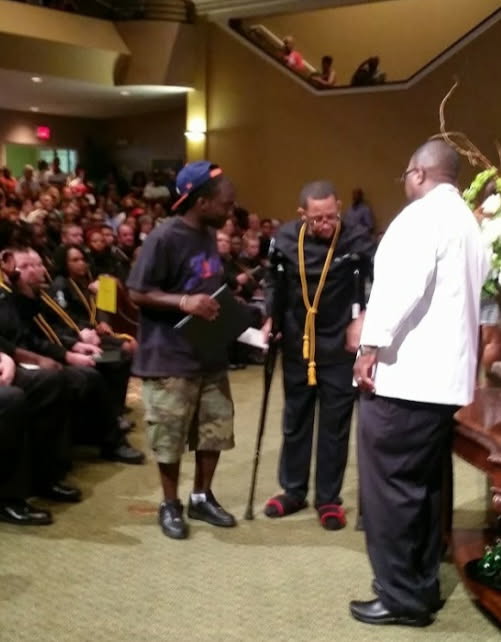 A man in graduation robes and a gold cord walking across a stage with a cane